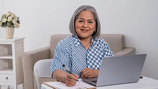 female business owner with gray hair sitting at table with laptop and notebook reviewing flood insurance policy in living room