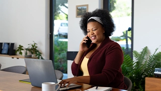 black woman talking on phone at table looking at personal insurance information on laptop